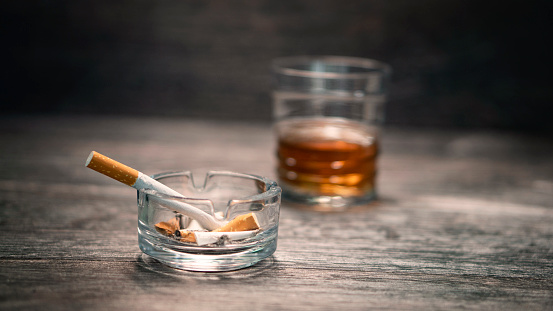 Small glass ash-tray with smoking cigarette and glass of whiskey on wooden background