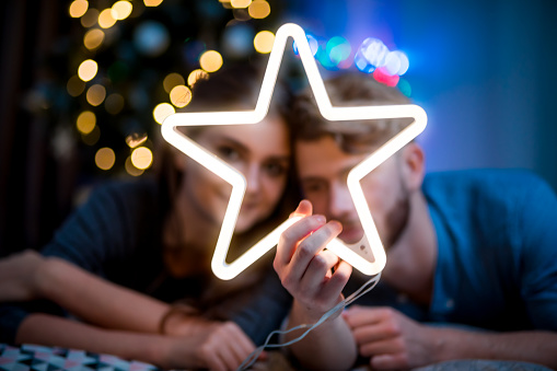 Couple in love at Christmas evening holding glowing white LED star, cozy dark living room