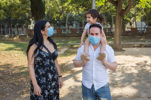 a caucasian father holds his 2 year old mestizo girl in a park in barcelona ci while the latin mother looks at them. the parents wear a protective mask against coronavirus. the girl looks smiling at the mother.