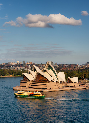 Sydney, Australia - August 2, 2019: A ferry crosses the waters of Circular Quay in Sydney. In the background looms the iconic form of the city's famous Opera House, beyond which we see the Royal Botanic Garden.
