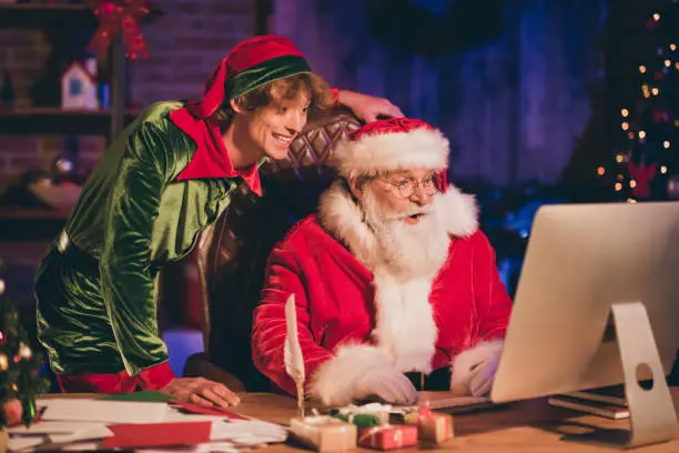 Photo portrait of santa claus and elf browsing internet on desktop.