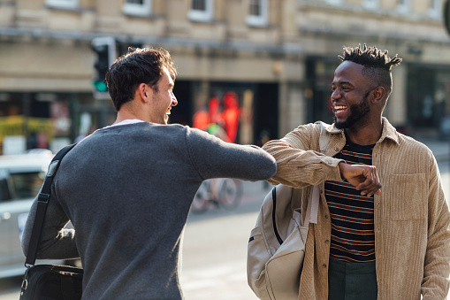 Two happy young male friends greeting each other with the elbow during the Coronavirus Covid-19 pandemic. They are outdoor in the city.