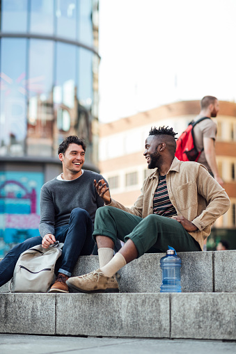 Two male friends are outdoors together chatting. They are relaxing sitting in the city center. They have a backpack and water bottle with them
