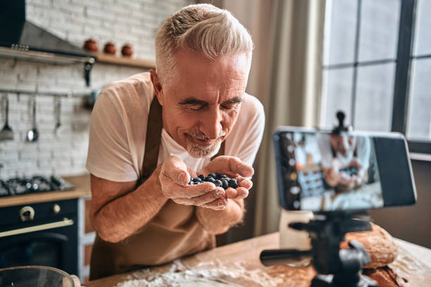 Adult chef sniffing aroma of fresh berries for baking Mature man holding berries in his hands over rolled dough for a pie in the kitchen while recording video on phone. Live streaming concept baking bread photos stock pictures, royalty-free photos & images