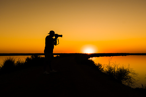 silhouette of a photographer with reflex camera and large lens in a lake taking pictures with the sunset in the background