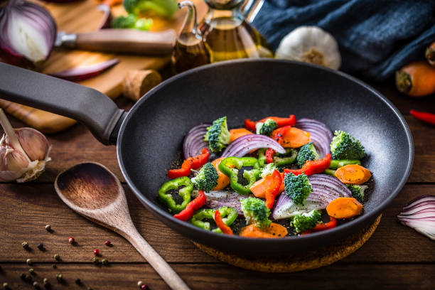 Top view of a cooking pan full of multicolored chopped vegetables ready to be stir fried. The composition includes chopped carrots, Spanish onions, broccoli and bell peppers. The cooking pan is at the center of the image and it's surrounded by a spatula and some vegan ingredients. Studio shot taken with Canon EOS 6D Mark II and Canon EF 100 mm f/ 2.8