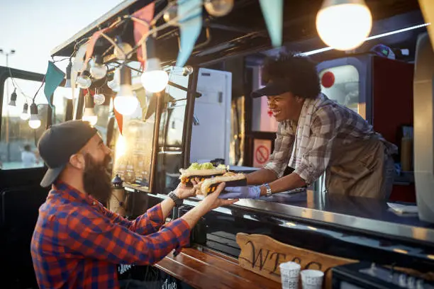 young caucasian beardy hipster taking two sandwiches from polite female afro-american employee in fast food service