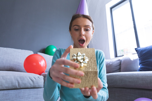 Caucasian woman at home celebrating birthday, in living room wearing party hat, gasping in surprise and opening a present. Social distancing during Covid 19 Coronavirus quarantine lockdown.