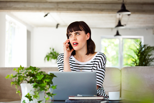 Young woman wearing striped t-shirt sitting on sofa in the creative workplace over laptop and talking on smart phone.