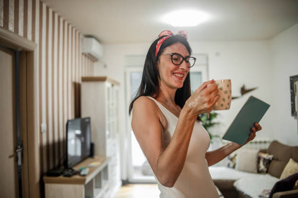 A young-looking senior woman reads a book to her apartment and holds a cup of tea - He uses the non-working day to relax with a book and a cup of tea una mujer mayor de pelo negro con gafas está descansando y caminando alrededor de su casa caliente leyendo un libro - una mujer mayor de aspecto joven lee un libro a su apartamento y sostiene una taza de té - utiliza el día no laborable para relajarse c