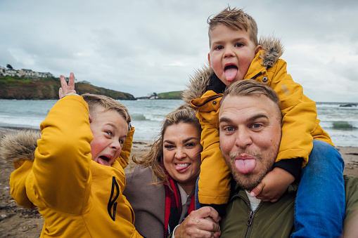 A selfie shot of a mid adult caucasian couple and their two young sons wearing casual outdoor clothing on a beach. The father has one of the boys on his shoulders while the mother is carrying their other son. They are all sticking their tongues out and pulling silly faces on an overcast autumn day at the beach. They are on a staycation to lower their carbon footprint and support the local economy.