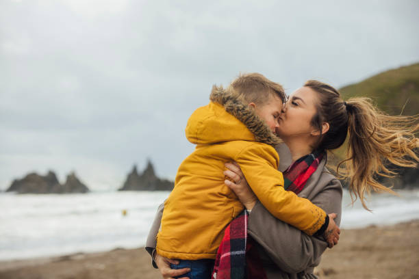 Beach Kisses A mid adult caucasian woman and her young son wearing casual outdoor clothing on a beach. The woman is carrying her son and kissing his head while he embraces her. It is an overcast autumn day in Devon. They are on a staycation to lower their carbon footprint and support the local economy. staycation photos stock pictures, royalty-free photos & images
