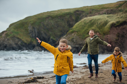 A mid adult caucasian man and his two young sons wearing casual outdoor clothing on a beach. They are all running along the beach with their arms up like an airplane on an overcast autumn day at the beach. They are on a staycation to lower their carbon footprint and support the local economy.