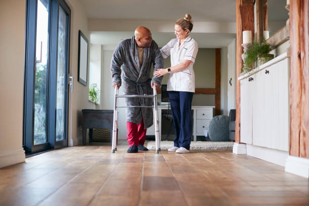 homme aîné dans la robe de chambre utilisant le cadre de marche étant aidé par le travailleur de soin féminin - healthcare worker home caregiver senior adult care photos et images de collection