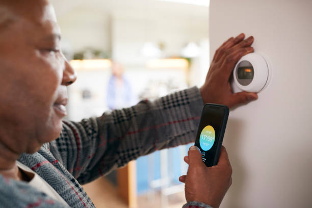 Mature Man Using App On Phone To Control Digital Central Heating Thermostat At Home Mature Man Using App On Phone To Control Digital Central Heating Thermostat At Home smart thermostat stock pictures, royalty-free photos & images