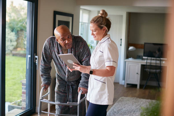 homme aîné dans la robe de chambre utilisant le cadre de marche étant aidé par l’infirmière femelle avec la tablette numérique - home caregiver senior adult community outreach nursing home photos et images de collection