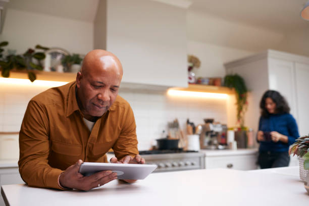 Mature Man At Home In Kitchen Looking At Digital Tablet With Female Partner In Background Mature Man At Home In Kitchen Looking At Digital Tablet With Female Partner In Background using digital tablet stock pictures, royalty-free photos & images