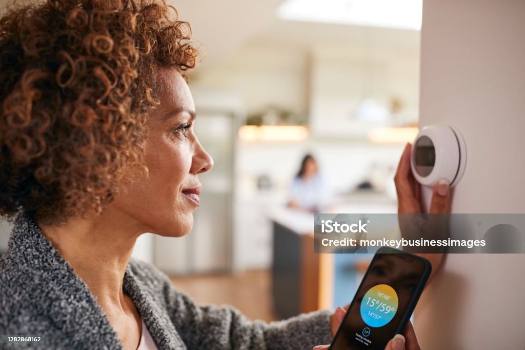 Mature Woman Using App On Phone To Control Digital Central Heating Thermostat At Home Thermostat Stock Photo