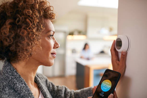 mulher madura usando aplicativo no telefone para controlar termostato de aquecimento central digital em casa - domótica - fotografias e filmes do acervo