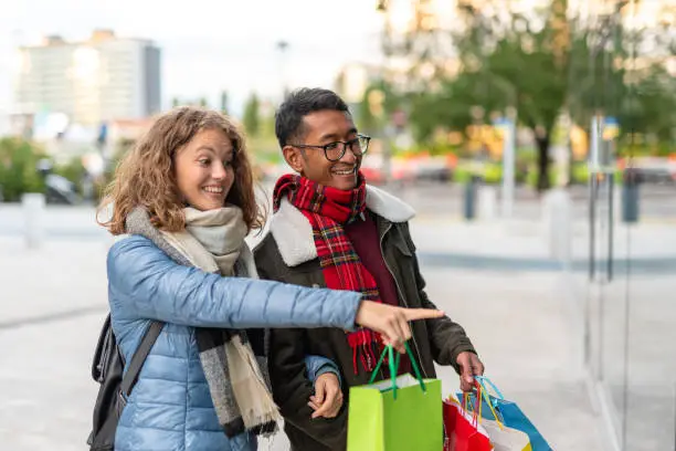 Photo of mixed couple at the time of shopping, young beautiful Caucasian woman indicates her desired to the store window, the Filipino boy smiles, concept of consumerism for the holidays