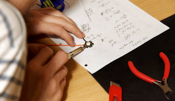 Young engineering college student soldering simple wired lambertian led diode on his desk, hands closeup, holding soldering iron. Temporary makeshift workspace table, mathematical notes, simple tools joven estudiante universitario de ingeniería soldar diodo lambertiano con cable simple en su escritorio, de cerca de las manos, sosteniendo soldador. tabla de espacio de trabajo improvisada temporal, notas matemáticas, herramientas sencillas
