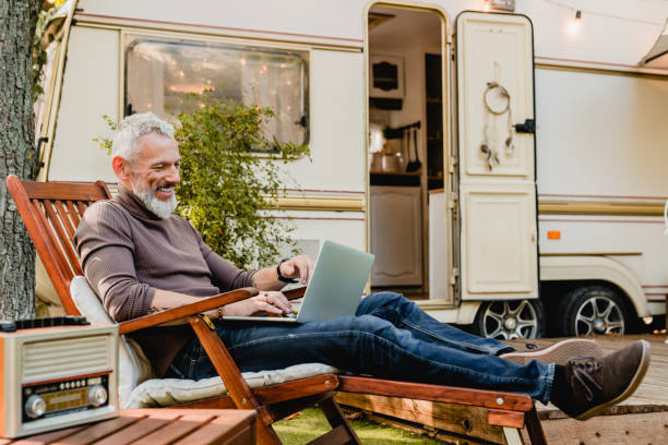 Attractive grey-haired man resting on the wooden deck chair using laptop with caravan van behind Attractive grey-haired man resting on the wooden deck chair using laptop with caravan van behind rv stock pictures, royalty-free photos & images