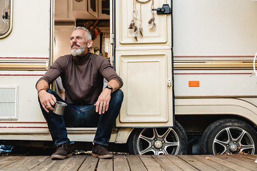 Portrait of handsome senior man with a beard sitting on the doorstep of the motor home holding iron mug
