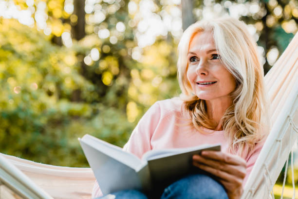 hermosa mujer rubia senior leyendo libro y sentado en la hamaca en el jardín - women book mature adult reading fotografías e imágenes de stock