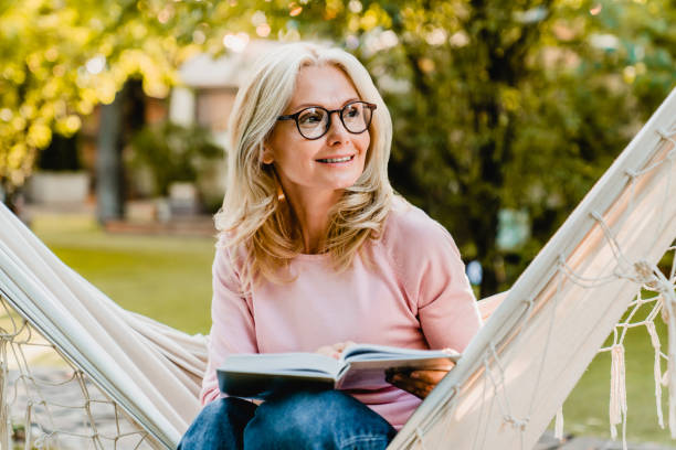 sonriente mujer rubia guapa de edad que lleva gafas mientras lee en la hamaca en el jardín de verano - women senior adult caucasian mature adult fotografías e imágenes de stock