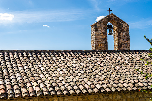 Partial view of a church in Provence. A very simple bell tower with an apparent bell and a cross. The roof is covered with Provencal clay tiles called canal tiles.
