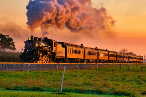 View of Golden Steam Passenger Train at Sunrise Traveling Thru Amish Countryside with lots of Smoke and Steam at Sunrise
