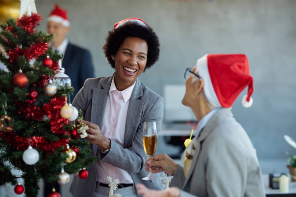 Cheerful black businesswoman and her colleague having fun while decorating Christmas tree in the office. Happy African American businesswoman decorating Christmas tree while talking to female colleague at office party. office christmas party stock pictures, royalty-free photos & images