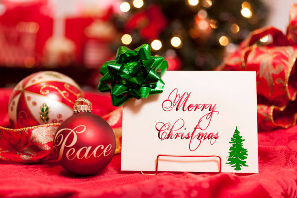 Holiday Greeting card with Christmas ornaments, and a Christmas tree with lights in the background.  Green gift bow, Merry Christmas and a small tree on the card.