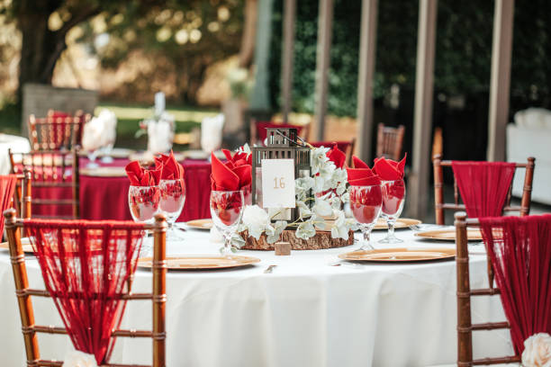 istock Wedding outdoor reception - round table with white table cloths, golden plates, rustic wooden centerpieces and black candle lanterns, eucalyptus leaves and red napkins. 1282899427