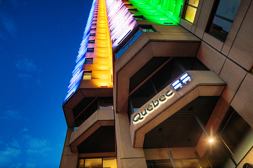 Quebec Government office tower in Downtown Montreal illuminated in a multi-colored pattern. The image is photographed at twilight and features the government logo and Quebec flag in the foreground.