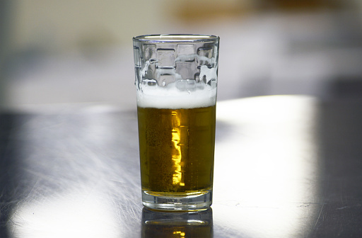 A golden pint of beer with foam at the top of the glass.  The glass of beer is placed on a hardwood surface with liquid drops of beer surrounding the glass on the table.