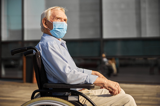 Side view of elderly man in a protective mask sitting in a wheelchair and enjoying the warm sunny day
