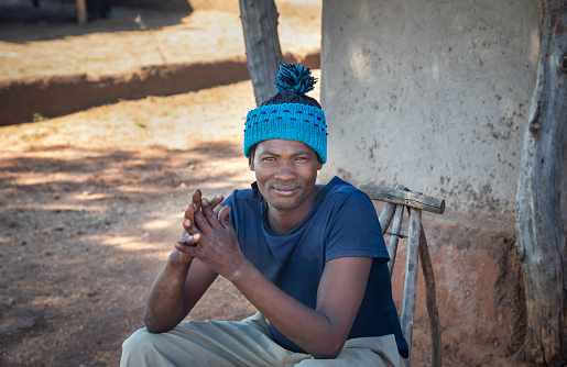 young african man with blue beanie sitting down on a wooden chair, village in Botswana