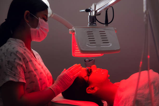 молодая женщина, имеющие красный светодиодный свет лицевой терапии лечения в салоне красоты. косметолог в маске для лица, поддерживая проц� - поддерживая стоковые фото и изображения