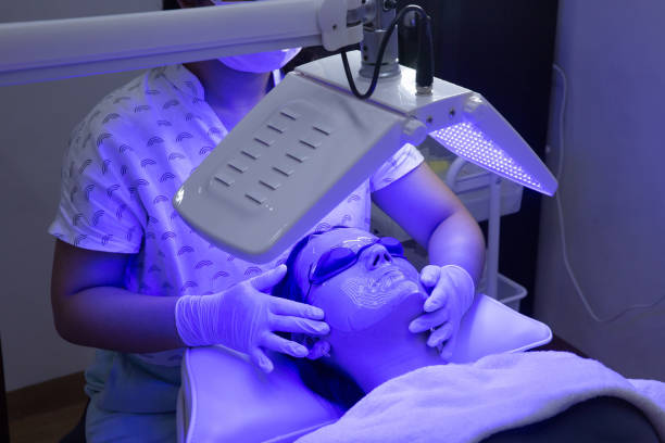 Young woman having blue LED light facial therapy treatment in beauty salon. Beautician wearing face mask maintaining safety procedures during appointment. Beauty, new normal and wellness Young woman having blue LED light facial therapy treatment in beauty salon. Beautician wearing face mask maintaining safety procedures during appointment. Beauty, new normal and wellness concept light therapy stock pictures, royalty-free photos & images