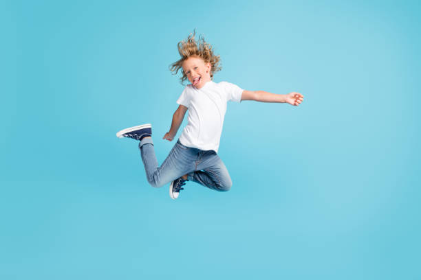 Photo of little child jump raise legs stick-out tongue wear white shirt jeans sneakers isolated blue color background Photo of little child jump raise legs stick-out tongue wear white shirt jeans, sneakers isolated blue color background t shirt caucasian photography color image stock pictures, royalty-free photos & images