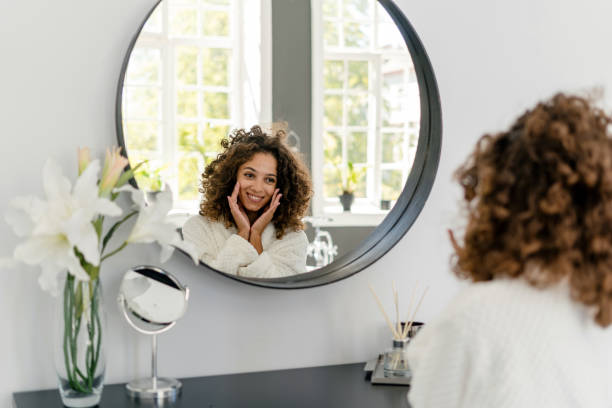 Young afro american woman in bathrobe at bathroom Natural beauty concept. Happy young afro american woman sitting in bathrobe near mirror at bathroom, smiling wide, enjoying morning routine, rejuvenation, face massage and spa procedure at home looking in mirror stock pictures, royalty-free photos & images