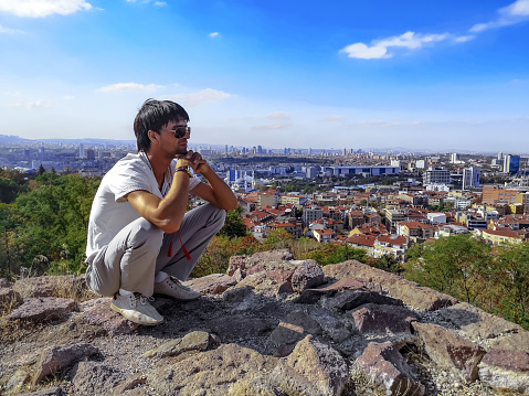 A young adult man in sunglasses is squatting on a hilltop in the center of Ankara overlooking the capital of Turkey. Tourist pensively looks at the city