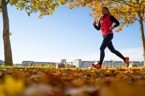 side low angle view of sporty happy smiling mature woman running outdoors on sunny autumn day with urban background