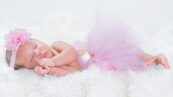 Cute newborn baby wears a pink flower crown lies swaddled in a white blanket.