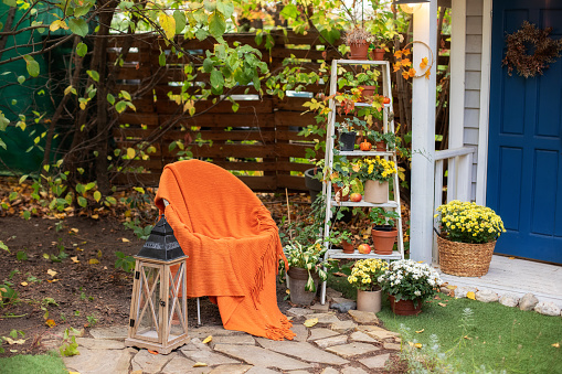 Cozy Autumn patio with chair, plaid, wooden lantern, potted chrysanthemums and pumpkins. Halloween. Decorations in backyard for relax in autumn garden. Stylish fall decor on front porch home for relax