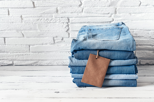 A stack of blue jeans with a brown blank tag on a white wooden table, brick backing. Heap of stylish trendy denim pants. Selective focus