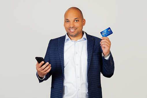 Portrait of happy young African American guy businessman using credit card to pay online using smartphone. Standing on a gray background