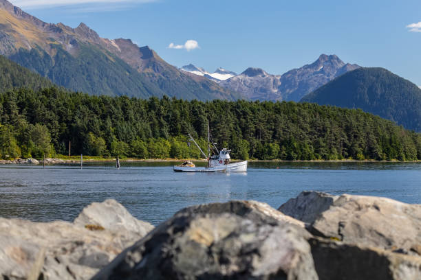 fishing boat drifting in harbor. rocks in soft focus in the foreground. mountains and forest in the background. sitka, alaska, usa - sitka imagens e fotografias de stock