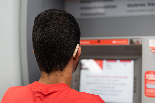 Deaf young man using hearing aid at bank ATM